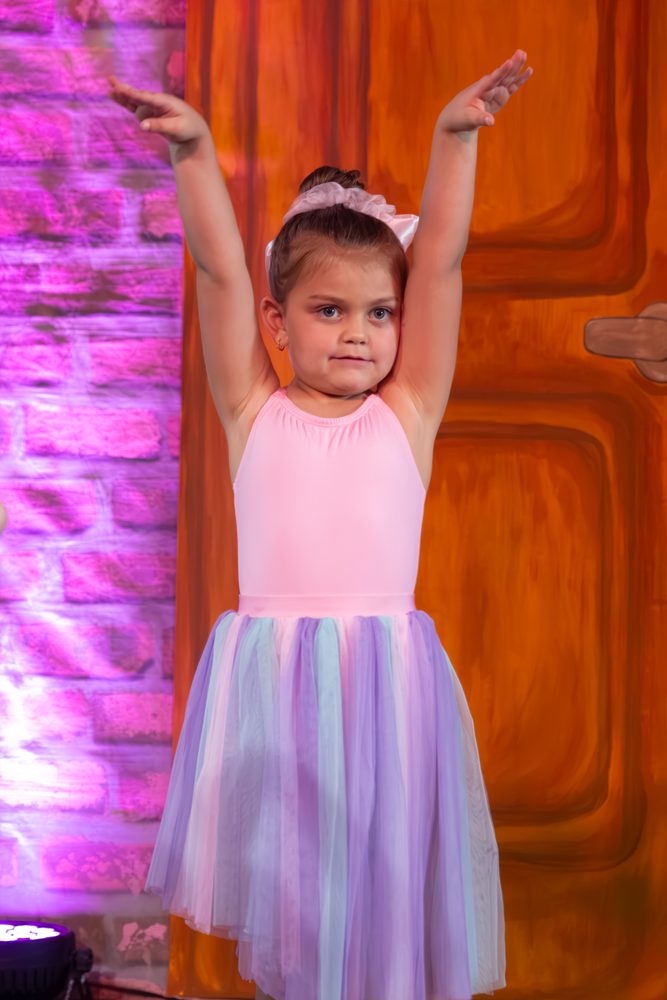 A girl in a pink leotard and pastel tutu with both arms raised on stage at a school concert