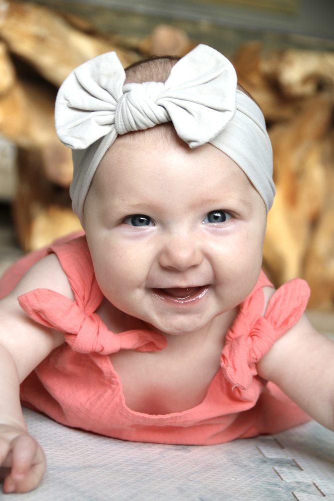 A smiling five-month-old baby in a coral top and grey bow headband