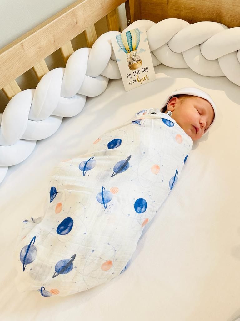 A newborn swaddled in a planet-print wrap, asleep in a cot