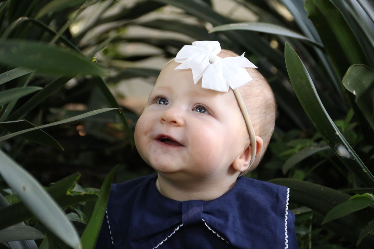 A smiling baby with a white bow peeking out between garden leaves