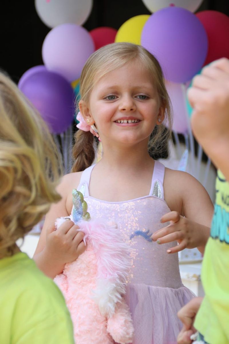 A smiling girl in a sparkly unicorn outfit holding a fluffy unicorn toy in front of balloons