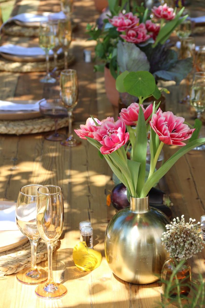 A long wooden party table with pink tulips, a brass vase and glasses in dappled sunlight