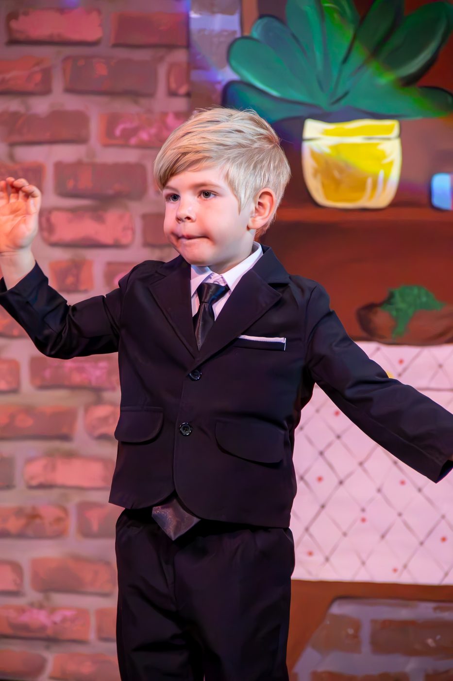 A young boy in a black suit and tie performing on stage in front of a painted brick-wall set