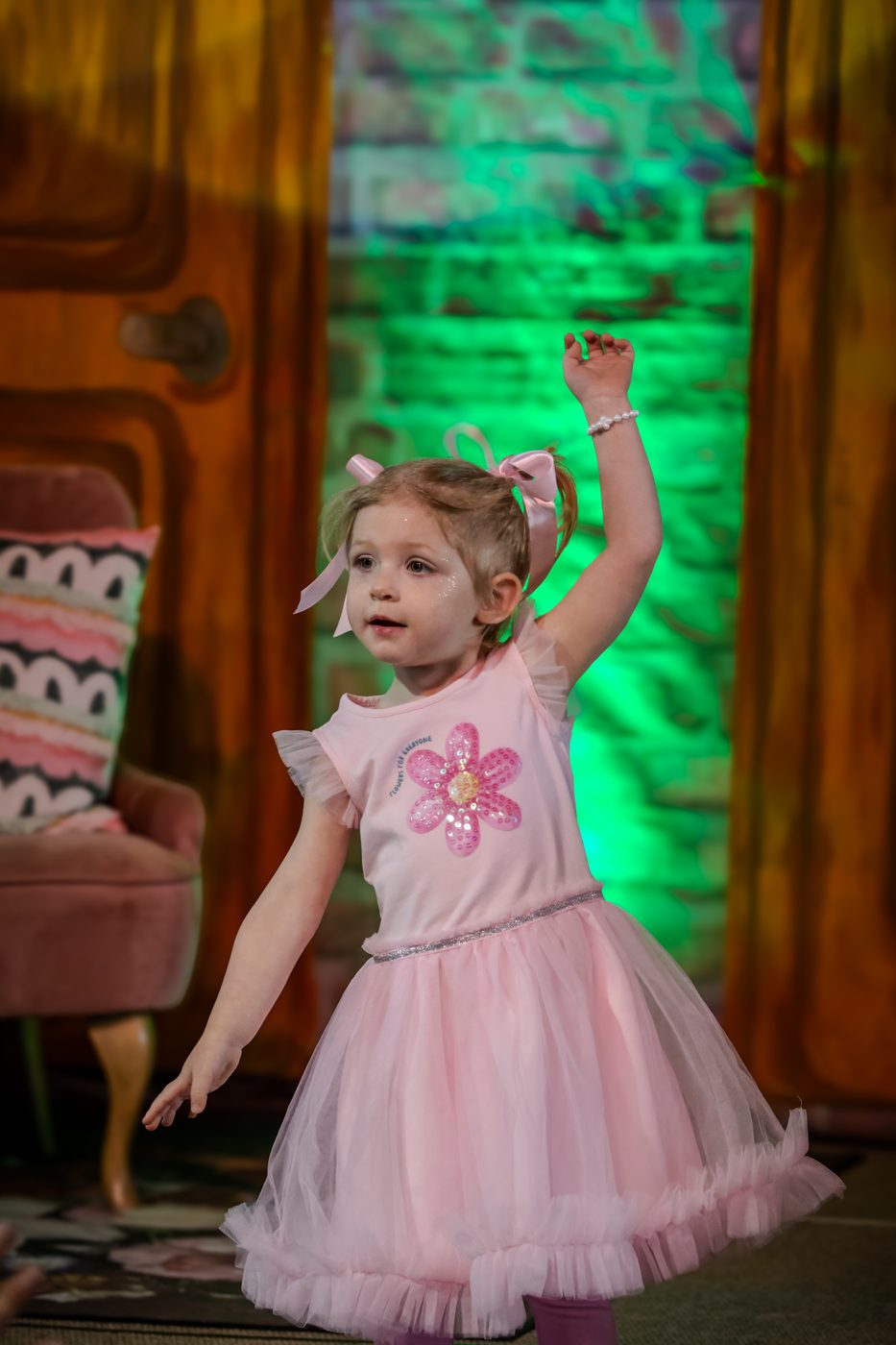 A little girl in a pink tutu dress dancing on stage with one arm raised under green stage lights