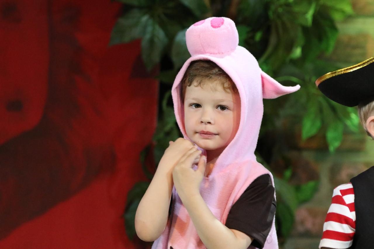 A young boy in a pink pig costume on stage beside a pirate, in front of a leafy set