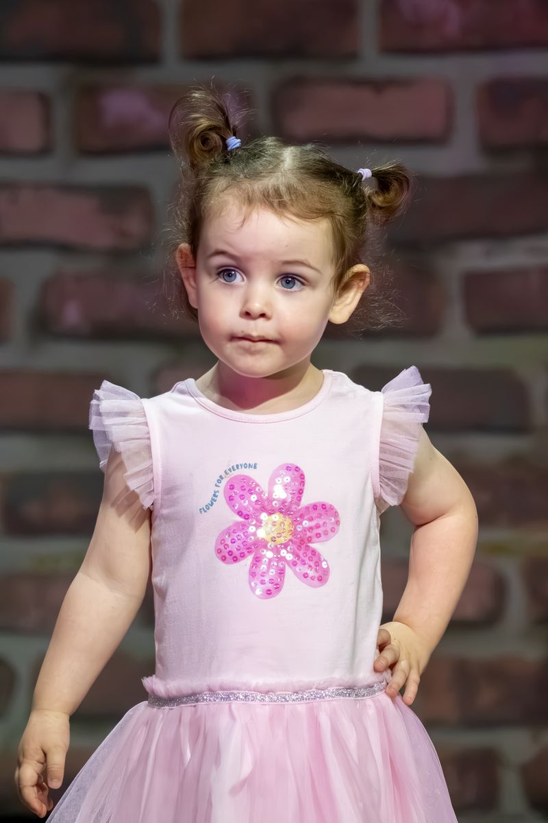 A little girl in a pink flower dress with a hand on her hip on stage